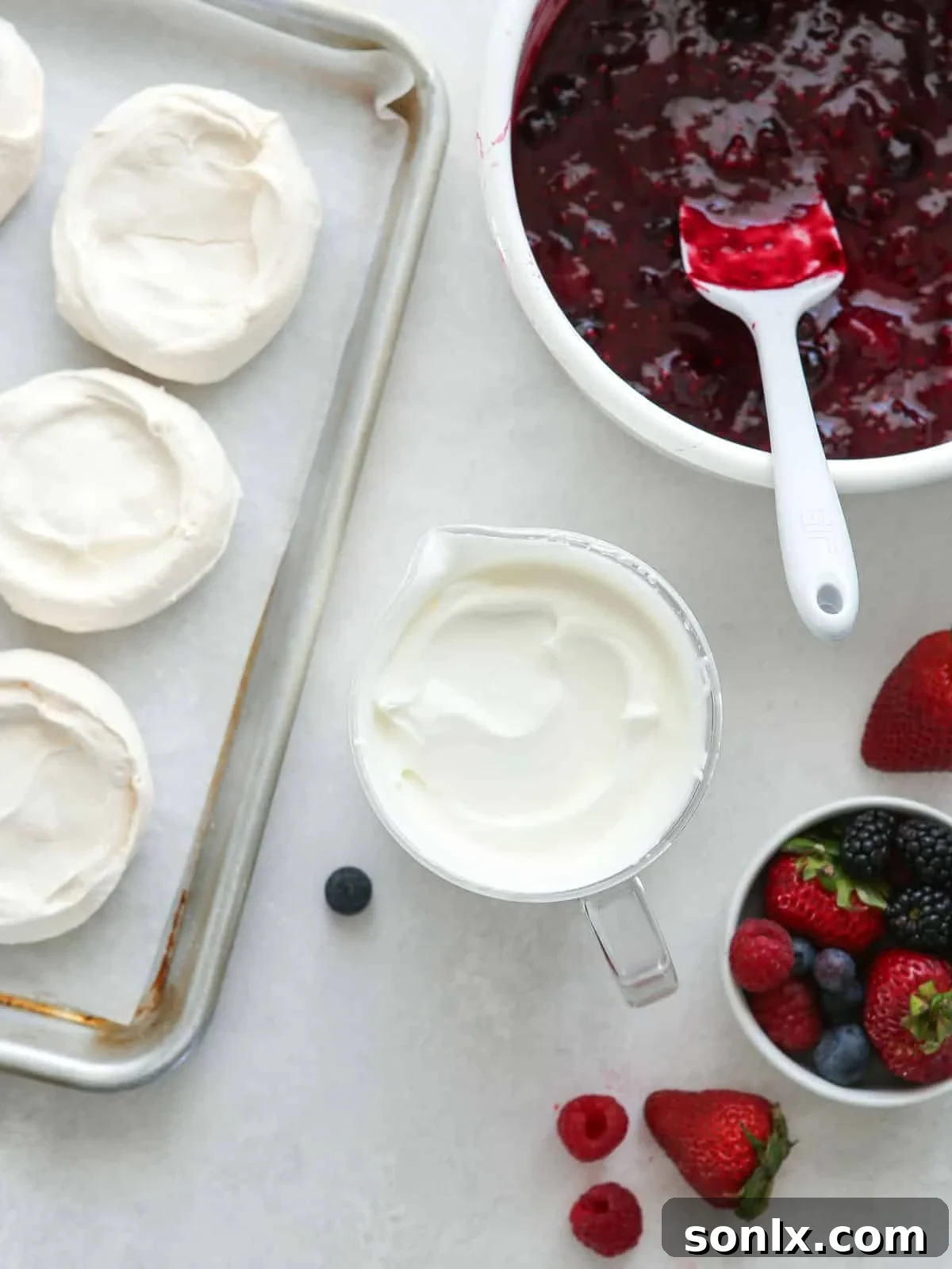 Mini baked pavlovas on a sheet pan, with sweetened whipped cream in a bowl and berry compote in a saucepan